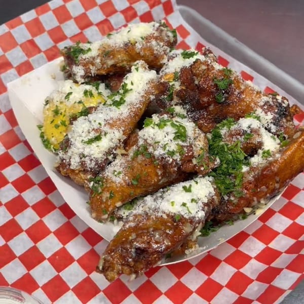 Crispy chicken wings topped with grated cheese and fresh parsley, served in a paper boat on red and white checkered paper.