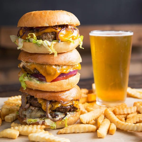 A towering stack of three cheeseburgers with lettuce, onion, and tomato, next to a glass of beer and crinkle-cut fries.