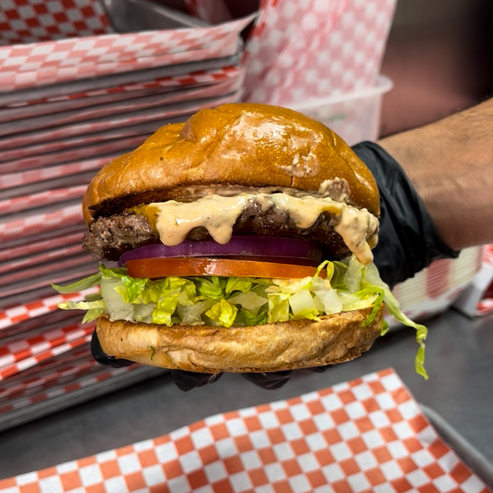 Juicy cheeseburger with lettuce, tomato, red onion, creamy sauce, and a toasted bun, held over red-and-white checkered paper.