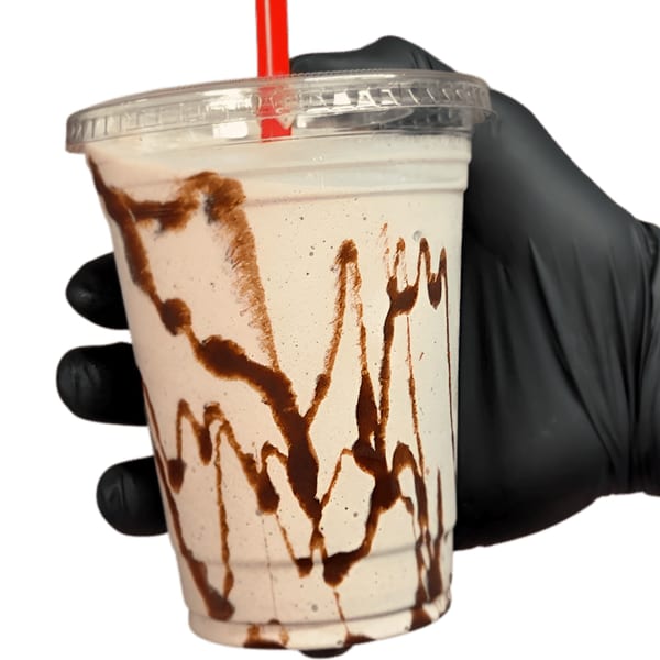 Hand in a black glove holding a milkshake with chocolate syrup streaks in a clear cup.