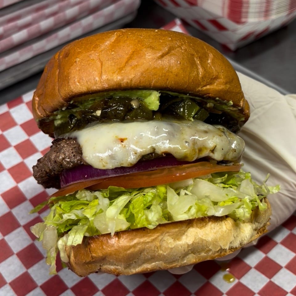 Cheeseburger with a toasted bun, melted cheese, grilled onions, tomato, lettuce, and pickles on red-and-white checkered paper.