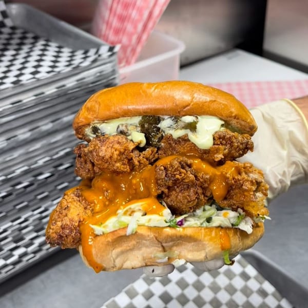A hand in a white glove holds a large fried chicken sandwich with coleslaw and sauces.