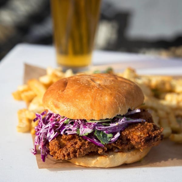 Crispy fried chicken sandwich with purple cabbage slaw and cilantro, served with fries and a beer.